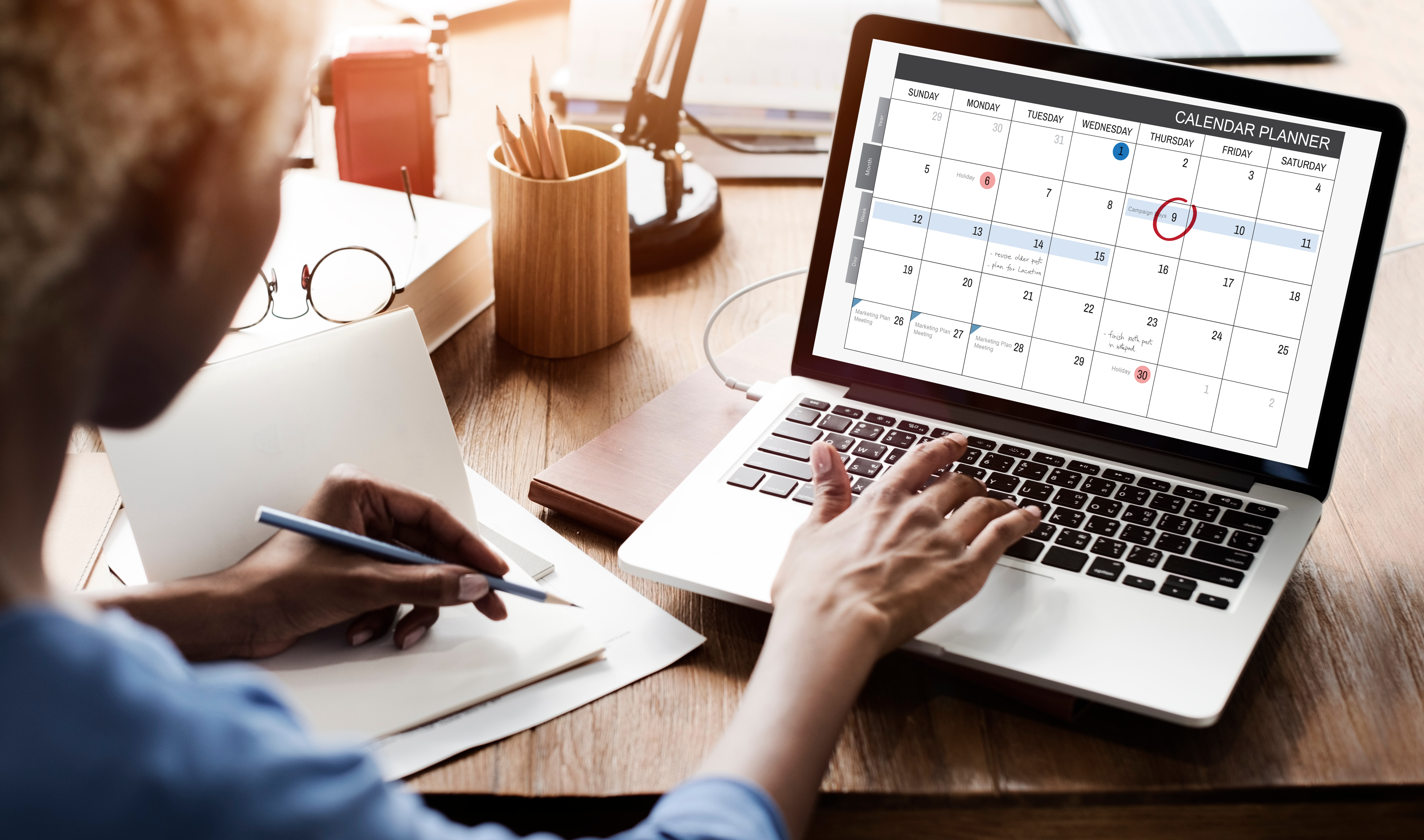 Alt tag: "A person planning their schedule using a calendar planner on a laptop screen, with notes and office supplies on a wooden desk in a well-lit room.